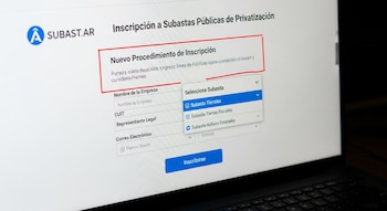 Una pantalla de laptop muestra el sitio web SUBAST.AR con un formulario de inscripción a subastas públicas y un menú desplegable con opciones de subastas.
