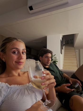 Una mujer sostiene una copa de vino en primer plano mientras un hombre sentado en un sofá sostiene un teléfono móvil junto a ella