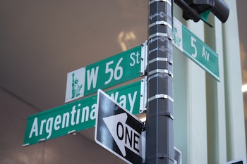 Poste con tres señales de calle verdes y una señal de dirección de un sentido. Las señales dicen W 56 St, 5 Av y Argentina Way. La de W 56 St tiene la Estatua de la Libertad