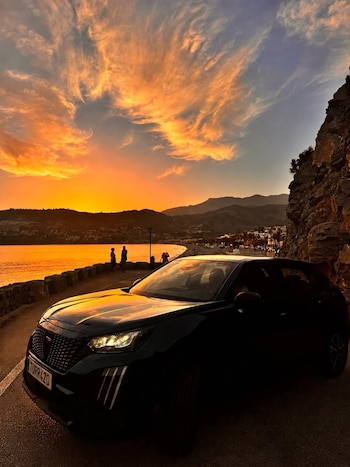 Coche oscuro en carretera costera al atardecer. Sol naranja se refleja en el mar y el cielo, con nubes vibrantes, sobre montañas y una playa