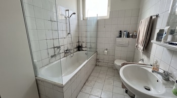 Interior de un baño con azulejos blancos y moho negro en las juntas, bañera, ducha, inodoro, lavabo, toalla y artículos de higiene.
