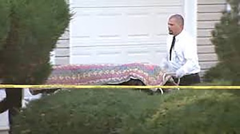 Un hombre en camisa blanca y corbata negra empuja una camilla con un cuerpo cubierto por una manta de colores frente a un garaje blanco, detrás de cinta amarilla de la policía