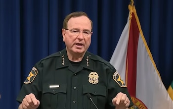 Hombre en uniforme de sheriff con gafas y micrófono, parado frente a una cortina azul y la bandera del estado de Florida