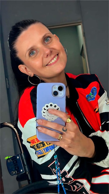 Laura Scianca, una mujer con cabello oscuro recogido y ojos azules, sonríe a la cámara mientras se toma una selfie. Viste una chaqueta colorida y se ve parte de su silla de ruedas