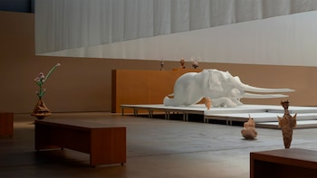 Una sala de exposición con una gran escultura de mamut blanco acostado sobre una plataforma. A su alrededor hay varias esculturas orgánicas y una planta con flores