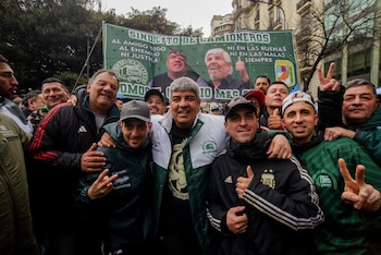 Dos hombres adultos, uno con chaqueta verde, el otro con chaqueta blanca y verde con logo sindical. Multitud y pancarta verde con retratos de fondo