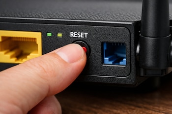 Mano presionando un botón rojo de reinicio en un router Wi-Fi negro, con puertos de red amarillos y azules, y luces indicadoras verdes.