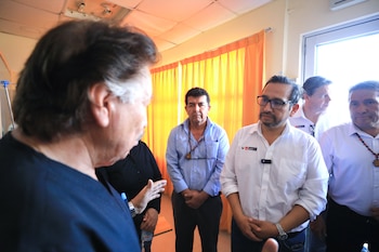 Un grupo de hombres, varios con camisa blanca y logo del Ministerio de Salud, escucha a un interlocutor dentro de una sala iluminada