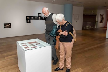 Un hombre y una mujer mayores observan objetos en una vitrina de cristal en una galería de arte con paredes blancas y suelo de madera. En el fondo se ven fotos enmarcadas