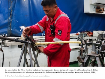 Hombre con overol rojo trabaja con herramientas en un cable grueso apoyado sobre dos soportes metálicos, con una funda amarilla en un extremo