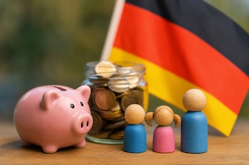 Una alcancía rosa con forma de cerdo, un frasco de vidrio con monedas de euro, tres figuras de madera pintadas y una bandera de Alemania.