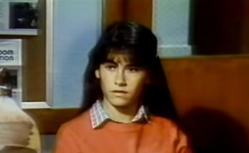 Retrato de una joven con suéter naranja, camisa a cuadros y cabello largo mirando hacia el frente en una habitación