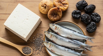 Un bloque de tofu, tres sardinas en un plato, semillas de chía, una cuchara de madera, tres higos secos y seis ciruelas pasas sobre una mesa de madera.