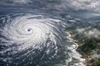 Un gran huracán con ojo central visible sobre el océano, nubes oscuras con lluvia y relámpagos, y una costa montañosa con vegetación.
