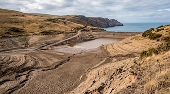 Terreno de barro seco y agrietado en un embalse con poca agua, rodeado de colinas secas y un acantilado junto al mar bajo un cielo nublado.