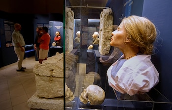 Mujer de perfil en primer plano. Vitrina con busto, cráneo humano y pilar de piedra. Personas y paneles informativos en segundo plano