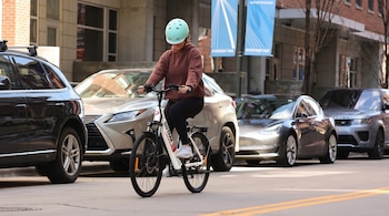 Adolescente en bicicleta eléctrica blanca con casco turquesa, moviéndose en una calle urbana flanqueada por varios autos estacionados y edificios al fondo.