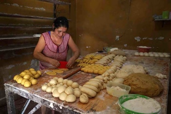 Mujer con delantal moldea masa sobre una mesa de madera. Hay varias porciones de masa de pan y recipientes con harina