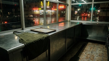 Delantal verde oliva y bandeja metálica sobre un mostrador de acero en un local con ventanales que dan a la calle de noche.