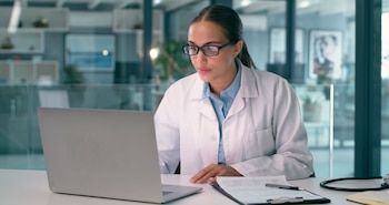 Una mujer con anteojos y bata blanca utiliza una computadora portátil sentada ante un escritorio con papeles y un estetoscopio