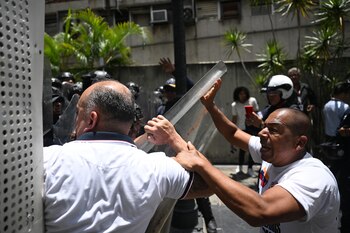 Dos hombres empujan un escudo transparente en primer plano. Detrás de ellos, varias personas con cascos y vegetación