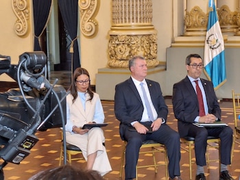 Tres personas sentadas: una mujer, un hombre y otro hombre. La mujer sostiene una tablet. Los hombres visten traje. Detrás, columnas ornamentadas y una bandera de Guatemala