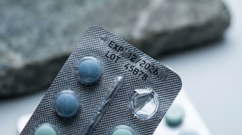 Primer plano macro de un blíster de pastillas azul acero con fecha de vencimiento 12/2026 y lote impresos en negro sobre el aluminio. Algunas pastillas faltan.