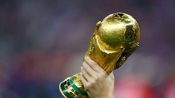 Primer plano de una mano sosteniendo el brillante trofeo de la Copa Mundial de la FIFA, de color dorado y con base verde, contra un fondo borroso