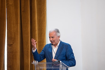 Un hombre de pelo blanco y traje azul gesticula mientras habla desde un podio transparente con un micrófono, frente a cortinas marrones y una pared blanca