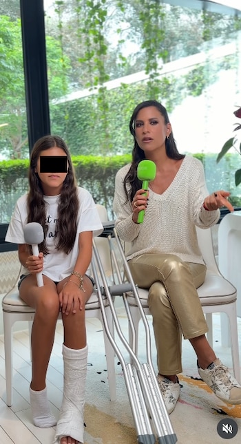 Dos mujeres sentadas en sillas frente a ventanas. Una joven tiene un yeso en la pierna izquierda y muletas al lado. Ambas sostienen micrófonos