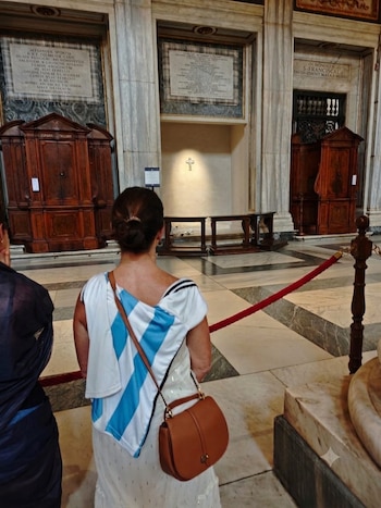 Mujer de espaldas, vestido blanco con rayas azules, cartera marrón, columnas de mármol, mobiliario de madera, cruz en la pared, cuerda roja, piso de mármol
