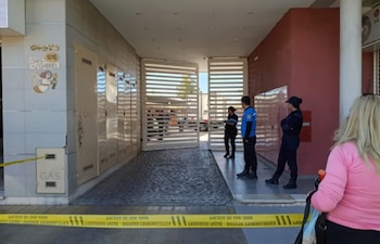 Policías uniformados vigilan la entrada de un edificio con puerta metálica y cinta policial amarilla. Una mujer con cabello rubio en primer plano