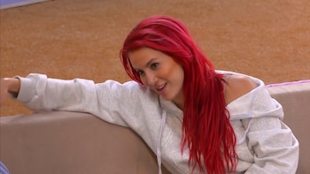 Mujer con cabello rojo y sudadera gris claro, recargada en un sofá color crema. Sonríe, lleva un piercing en la nariz y mira hacia su derecha