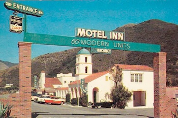 Entrada del Motel Inn con letrero, edificios de estilo colonial español con torre y cúpula. Varios coches de época en el estacionamiento. Fondo de montañas