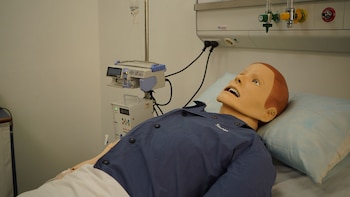 Maniquí médico acostado en una cama de hospital con suero, bombas de infusión y paneles de gas en la pared