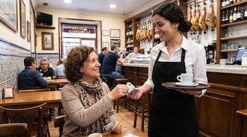 Una mujer con cabello rizado y bufanda de leopardo entrega billetes de euro a una camarera sonriente con delantal negro y bandeja con café. Fondo con jamones colgados.