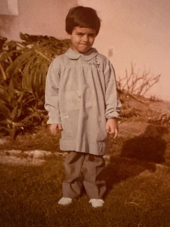 Un niño con guardapolvo claro y pantalón oscuro de pie sobre el pasto. Fondo con plantas y pared clara. La imagen tiene tonos sepia