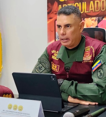 Hombre con uniforme militar verde y borgoña, parches de la Guardia Nacional Bolivariana, bandera de Venezuela, sentado en escritorio con tableta, micrófono, placa con nombre