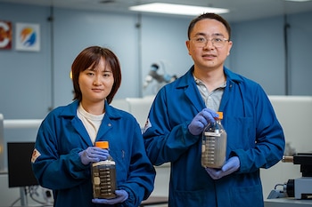 Una mujer y un hombre con batas de laboratorio azules y guantes sostienen frascos de vidrio con polvos, uno oscuro y otro claro, en un laboratorio