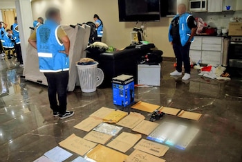 Oficiales de policía en chalecos azules inspeccionan un interior con documentos vehiculares, placas y sobres con nombres de autos esparcidos en el suelo