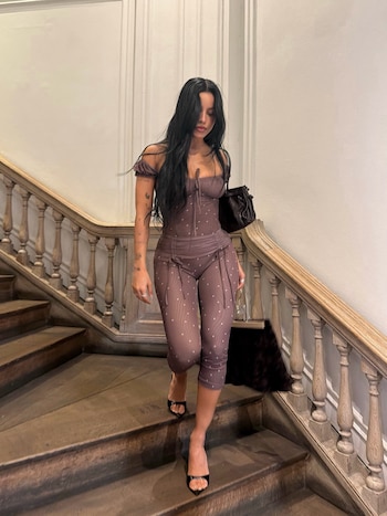 Emilia Mernes desciende por una escalera de madera con un conjunto ajustado de color marrón y un bolso negro