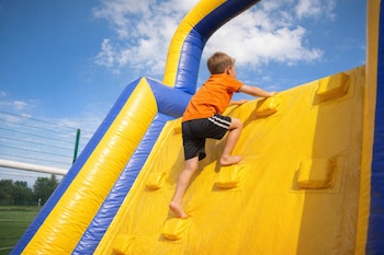 Un niño de aproximadamente siete años, con camiseta naranja y pantalones cortos negros, sube descalzo por la pared amarilla de un juego inflable azul y amarillo.