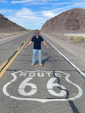 Hombre de pie en una carretera asfaltada frente al símbolo de la Ruta 66 pintado en el suelo entre cerros rocosos