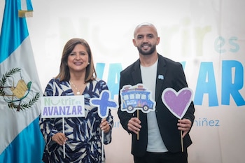 Dos personas sonrientes sostienen carteles con texto, un signo más, un autobús y un corazón. Detrás, la bandera de Guatemala y un banner