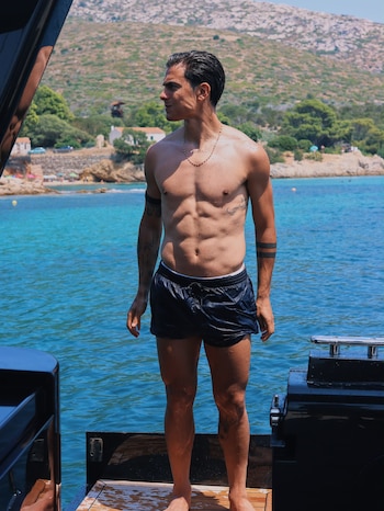 Hombre de pie sin camiseta y bañador oscuro en un barco. Al fondo, mar azul, costa con colinas, casas blancas y una playa de arena