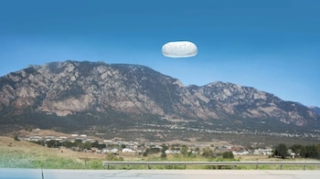 Imagen panorámica de un objeto blanco ovalado flotando sobre montañas rocosas, valles verdes y un pequeño pueblo bajo un cielo azul claro