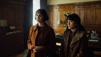 Dos mujeres con cabello oscuro y flequillo, vistiendo abrigos marrones, están de pie en una habitación con paneles de madera y luz tenue