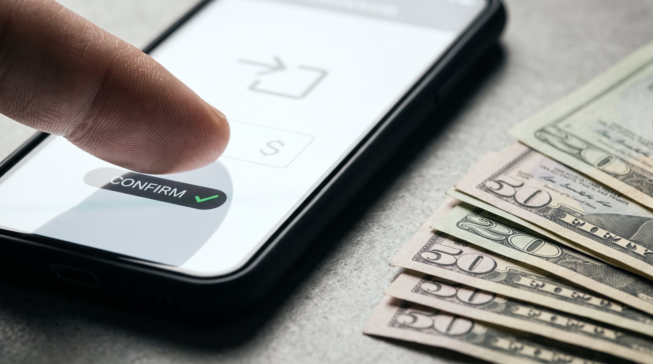 Un usuario confirma una transferencia bancaria desde su teléfono inteligente junto a varios billetes de dólares estadounidenses dispuestos sobre una mesa. (Imagen Ilustrativa Infobae)
