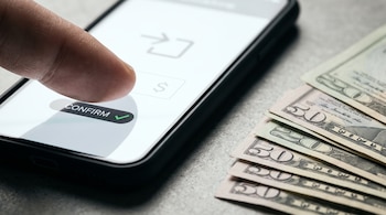 Un dedo pulgar sobre la pantalla de un teléfono con el botón confirmar, junto a billetes de veinte y cincuenta dólares sobre una superficie gris.