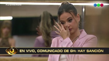 Una mujer con blazer rosa se cubre el rostro con la mano y llora. En la pantalla, un banner inferior anuncia una sanción de Gran Hermano